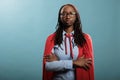 Brave and proud superhero woman wearing mighty hero cloak while standing with arms crossed on blue background.