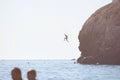 Brave young man jumping from high rocky mountain into t sea with swimming people during summer vacation