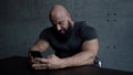 Brawny bald man is sitting at table at home and using smartphone, playing games or checking email