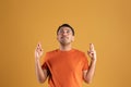 Brazilian man in orange t-shirt making cross fingers gesture and looking up, yellow background, studio shot, free space