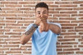 Brazilian young man standing over brick wall laughing at you, pointing finger to the camera with hand over mouth, shame expression