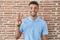 Brazilian young man standing over brick wall showing and pointing up with fingers number three while smiling confident and happy