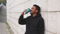 Break time! Happy smiling african man drinking coffee in black hoodie on city street over gray brick wall