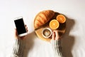 Breakfast in bed flat lay. Woman hands holding cup of coffee and phone, wooden cut with fresh croissant and strawberry in bed. Top