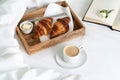 Breakfast in bed with two croissants and butter on the wooden tray, a cup of coffee and open book on the white sheet with blanket. Royalty Free Stock Photo