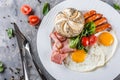 Breakfast, fried eggs, bun, sausage, bacon, prosciutto, fresh salad on plate on grey table surface. Healthy food, top view Royalty Free Stock Photo