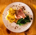 Breakfast meal plate of food comprising eggs Benedict with ham, spinach, toast and garnish set atop wooden table