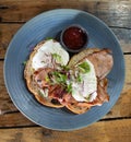 Breakfast meal plate of food comprising poached eggs with bacon, toast, sauce and garnish set atop wooden table