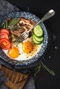 Breakfast set. Pan of fried eggs with bacon, fresh tomato, cucumber, sage and bread on dark serving board over black Royalty Free Stock Photo