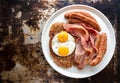 Breakfast on a white plate, chipolata sausage, bacon, fry eggs on top of hashbrowns, rustic tabletop view