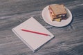 Breakfast at work. Tasty sandwich with sausage and cheese on white plate, notebooke with a pencil. Top view, vintage effect, gray