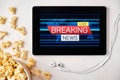 Breaking news logo on the screen of the tablet with popcorn box and Apple earphones on the background. Advertising or