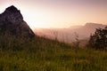 Breathtaking mountain landscape - bright pink golden sunset with sunlights, mist above lush green grass meadow, stone on slope