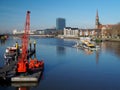 Bremen, Germany - February 14th, 2019 - Pier with several small vessels and bright red floating crane, barge with dredger and city