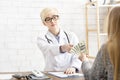 Bribery and corruption in medicine. Serious lady doctor in glasses and white coat takes dollars from patient