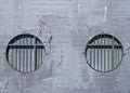 Brick wall with faded grey cracked peeling paint with two round blocked windows with green rusting metal bars