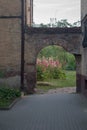 Brickwork arch connects corners of old buildings with shabby walls over tiled courtyard, behind growth of pink mallows.