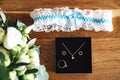 Bride accessories flat lay: black box with golden jewelry, white lace garter with light blue ribbon bow and white