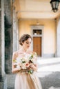 Bride in a beautiful dress with a bouquet of pink flowers stands in an arched corridor. Lake Como, Italy