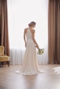 Bride in beautiful wedding dress with bouquet near window indoors, back view Royalty Free Stock Photo