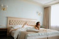 Bride with dark hair rest in wedding dress on bed. Bored young lady try to reach out to shoe on leg, mirror lying near. Royalty Free Stock Photo