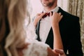 Bride fixes red bow tie on groom`s neck while they stand near the window