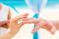 Bride giving an engagement ring to her groom under the arch decorated with flowers on the sandy beach. Wedding ceremony on