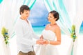 Bride giving an engagement ring to her groom under the arch decorated with flowers on the sandy beach. Wedding ceremony on