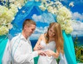 Bride giving an engagement ring to his groom under the arch decorated with flowers on the sandy beach. Wedding ceremony on