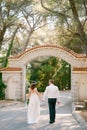 The bride and groom go to the beautiful arch at the entrance to the park holding hands, back view