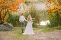 Bride and groom hold hands, go along the path to the water in autumn forest, girl with blond hair in a gray gorgeous