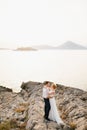 The bride and groom hug on the rocks by the sea against the backdrop of the mountains and the island of Mamula