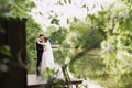 Bride and groom posing on the wooden pier near the pond among greenery. Young people embrace each other, and look at Royalty Free Stock Photo