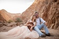 Bride and groom sit and smile in canyon on sand on background of rocks.