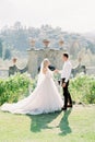 Bride and groom stand holding hands on the lawn near the stone wall with decorative vases Royalty Free Stock Photo