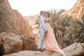 Bride and groom stand and kiss in canyon against background of rocks and palm trees.