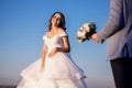 The bride and groom are standing near the river against the blue sky, the man carries a bouquet of white roses and gives his woman Royalty Free Stock Photo