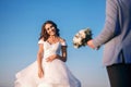 The bride and groom are standing near the river against the blue sky, the man carries a bouquet of white roses and gives his woman Royalty Free Stock Photo