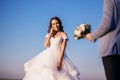 The bride and groom are standing near the river against the blue sky, the man carries a bouquet of white roses and gives his woman Royalty Free Stock Photo