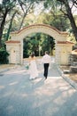 Bride and groom walk holding hands to the arched entrance to the park