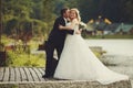 Bride holds groom's hand while he hugs her on the lake's shore Royalty Free Stock Photo