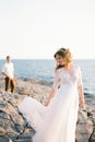 Bride lifts the hem of her dress on the rocks by the sea against the background of the groom Royalty Free Stock Photo