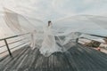 Bride in long white flying dress and veil standing over the ocean beach