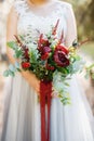 A bride in a pale gray wedding dress holds a bouquet with red peonies, astilba, roses and eringium and red long ribbons