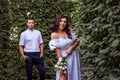 Bride playfully bared her shoulder and peeks out from behind the foliage with smile, groom standing and looking behind Royalty Free Stock Photo