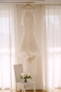 The bride`s dress by the curtains on the window above the chair with a bouquet of flowers. Royalty Free Stock Photo