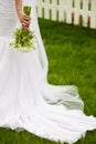 Bride`s hand with wedding bouquet of beige flowers over white wooden fence and green lawn. Trendy dress with vapoury veil. Royalty Free Stock Photo