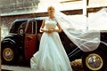 Bride's veil spreads over a retro car while she stands behind it