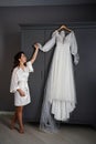 bride in a silk robe next to a white wedding dress hanging on the closet.