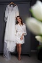 bride in a silk robe next to a white wedding dress hanging on the closet.
